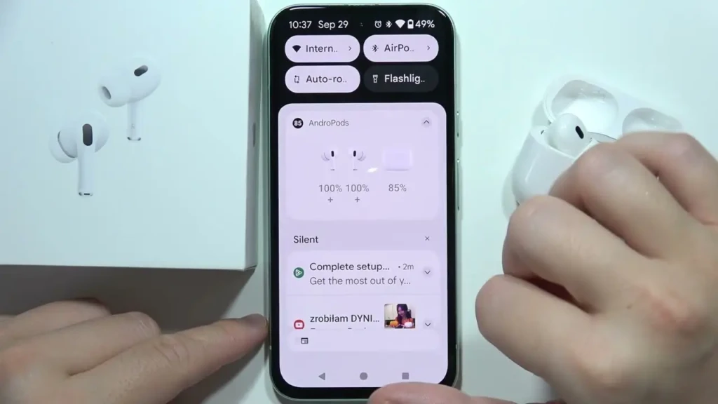 A smartphone screen displays an app interface for controlling earbuds, with battery levels shown. Nearby are the earbuds' case and packaging, suggesting tech usage.
