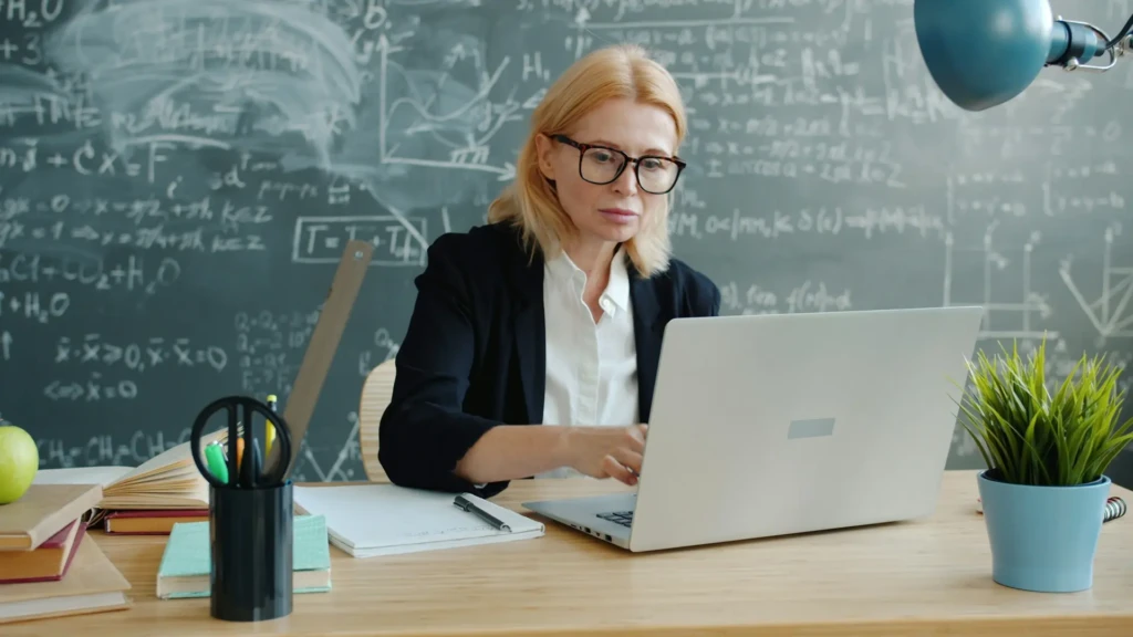 A focused female teacher with glasses working on a laptop at a wooden desk, with a chalkboard covered in math and chemistry equations behind her, plus books, stationery, an apple, and a small plant, depicting an academic environment blending traditional teaching with digital tools.