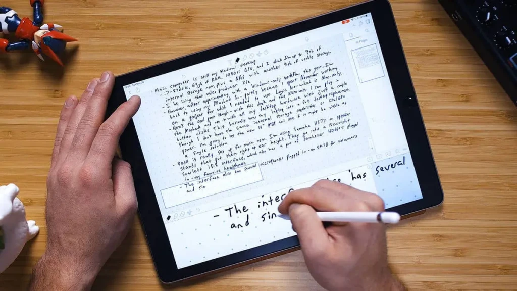 A person writing handwritten notes with an Apple Pencil on an iPad placed on a wooden desk, surrounded by small figurines; the screen shows dense, structured text in a dot-grid notebook layout, illustrating expressive, analog-style digital note-taking in a creative workspace.