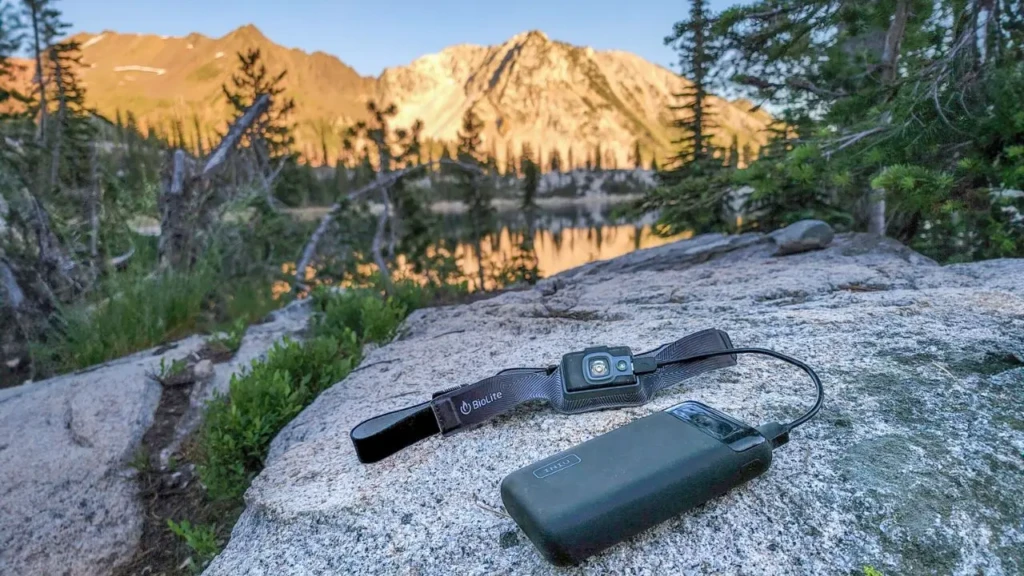 A rugged outdoor scene with a BioLite headlamp and a compact black power bank resting on a granite rock, set against a scenic mountain lake at golden hour, illustrating reliable portable power for camping and adventure use cases.