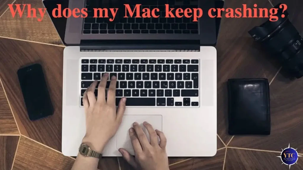A top-down photo of hands typing on a silver MacBook keyboard, with a smartphone, camera lens, and wallet nearby on a wooden desk, overlaid with the question “Why does my Mac keep crashing?” in bold red text, representing a real-world user experiencing repeated crashes and seeking answers.