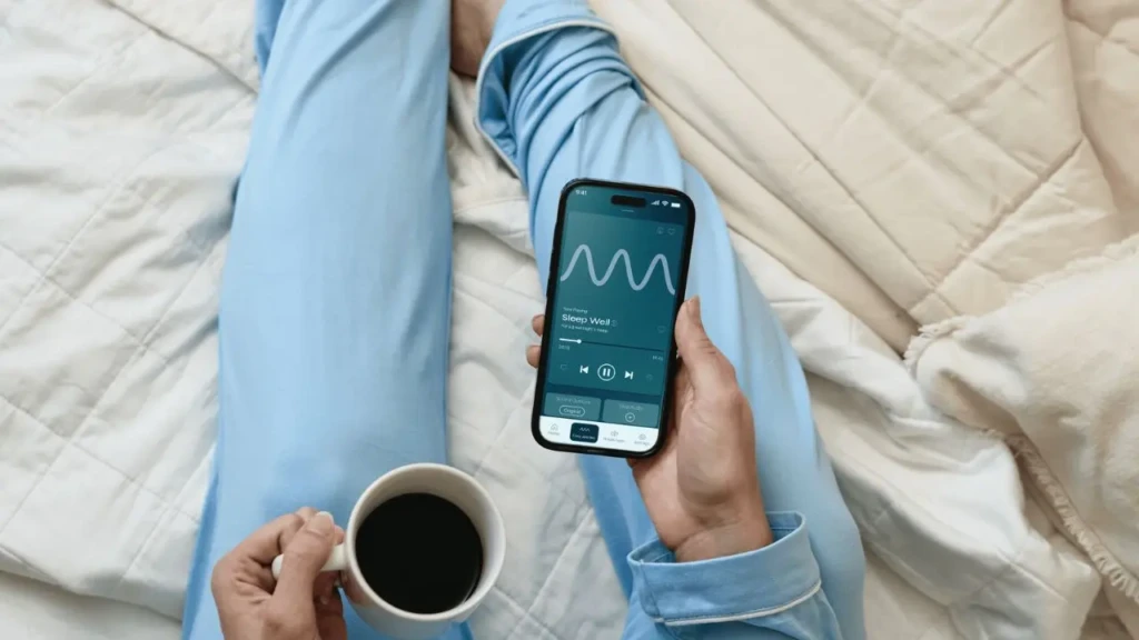 A person in light blue pajamas lying in bed, holding a smartphone displaying the “Sleep Well” audio session with a waveform visualizer, while their other hand holds a white mug of black coffee, illustrating a relaxed, pre-sleep or wind-down routine using a wellness app.