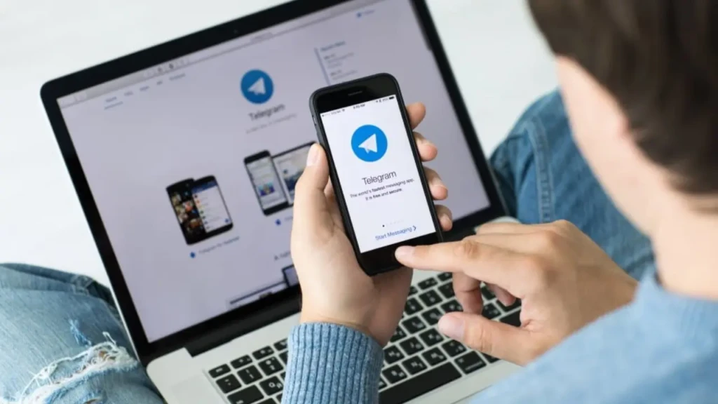 A person in jeans holds a smartphone showing the Telegram logo, with a laptop in the background also displaying the Telegram website, suggesting connectivity.