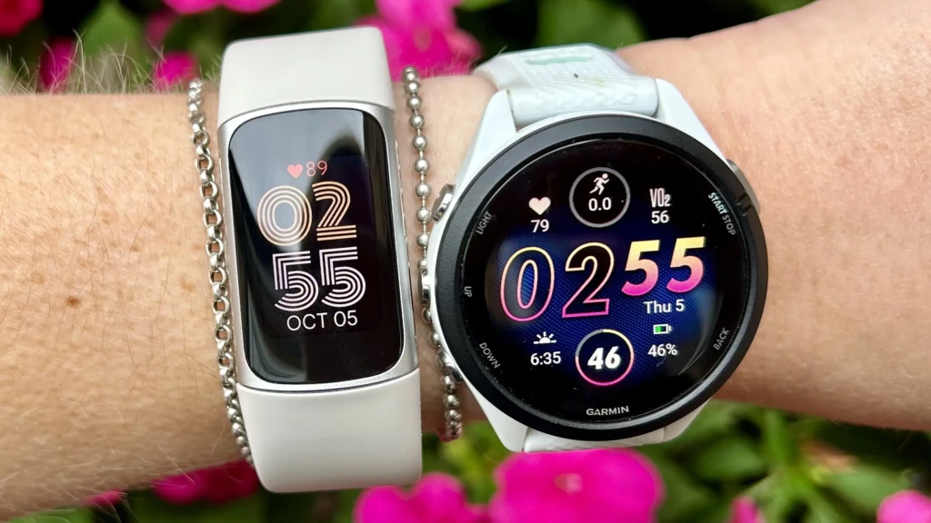Two wearables on a wrist against pink flowers: a white fitness band displaying “02:55 OCT 05” and heart rate 89, and a white Garmin smartwatch showing “02:55”, VO₂ 56, heart rate 79, and battery at 46%, demonstrating real-time health metrics and multi-sport functionality in an outdoor setting.