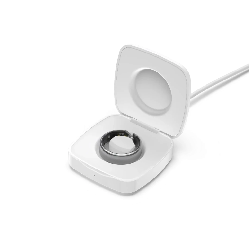 A white smart ring resting in an open square charging with a magnetic dock, connected to a charging cable, demonstrating wireless charging functionality and clean, modern accessory design.