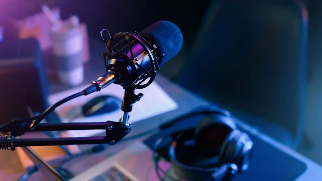 A dimly lit podcast studio setup with a microphone on a stand, headphones, and a laptop on a desk. Purple and blue hues create a focused, relaxed atmosphere.