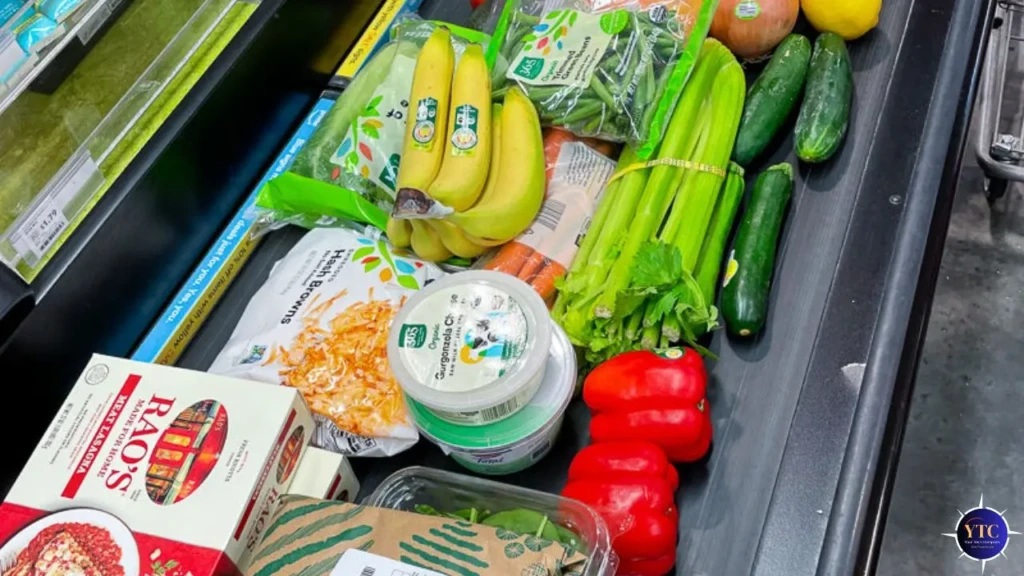 Grocery conveyor belt with bananas, celery, red bell peppers, cucumbers, spinach, tortilla chips, carrots, onion, coconut milk, and frozen cheese.
