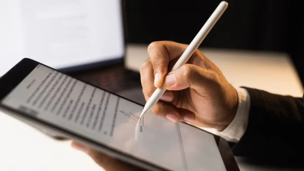 A hand holds a stylus and writes on a tablet, displaying text and graphics. The setting is a dimly lit workspace, conveying focus and technology.