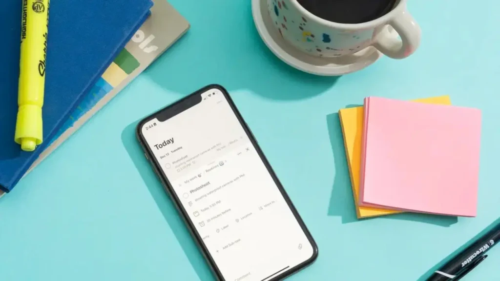 A smartphone on a teal desk displaying a “Today” task list, surrounded by physical productivity tools: a yellow highlighter, blue notebook, coffee mug, sticky notes, and a pen, evoking a hybrid analog-digital workflow for personal organization and daily planning.
