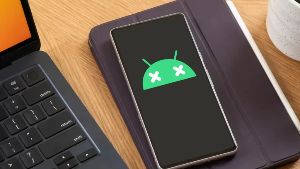A smartphone resting on a tablet case beside a laptop, displaying the Android robot logo with two white X marks over its eyes, symbolizing a crashed, frozen, or malfunctioning Android system, often used to represent boot loops or critical software errors.