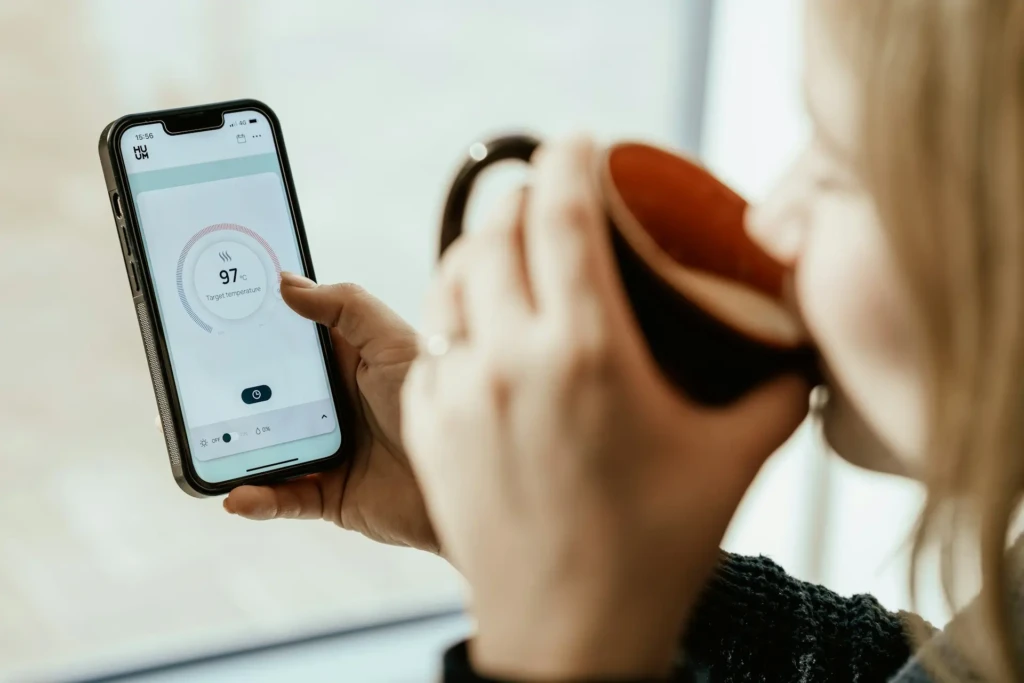 A person holding a smartphone displaying the HUUM sauna controller app, showing a target temperature of 97°C with a circular progress indicator, while sipping from a ceramic mug, illustrating smart home integration and relaxed user experience.