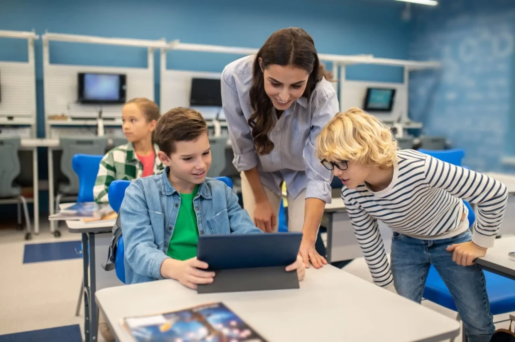 A teacher leans over a desk, smiling as two students, one holding a tablet and the other leaning in with curiosity, engage with digital learning in a modern classroom; a third student observes in the background, with blue chairs, mounted monitors, and a chalkboard visible,  illustrating collaborative, technology-enhanced instruction.