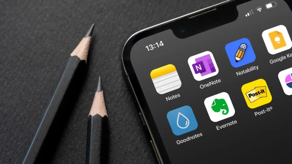 A close-up of an iPhone screen displaying six note-taking app icons, Notes, OneNote, Notability, Google Keep, Goodnotes, and Evernote alongside two sharpened black pencils on a dark textured surface, highlighting popular cross-platform note-taking tools in a creative workspace context.