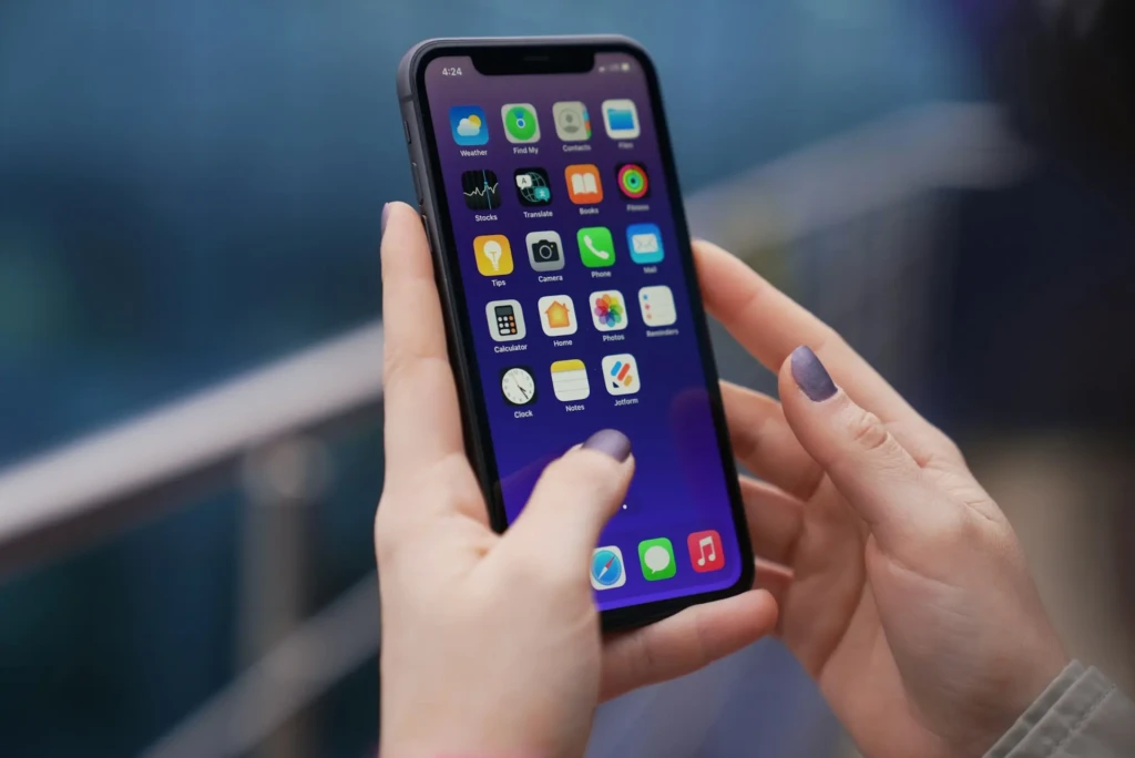 A pair of hands holding an iPhone with a vibrant purple home screen, navigating through app icons including Weather, Translate, Photos, Notes, and Jotform; the user’s thumb hovers over the screen, and nails are painted matte purple, highlighting everyday iOS interaction and personalized device use.