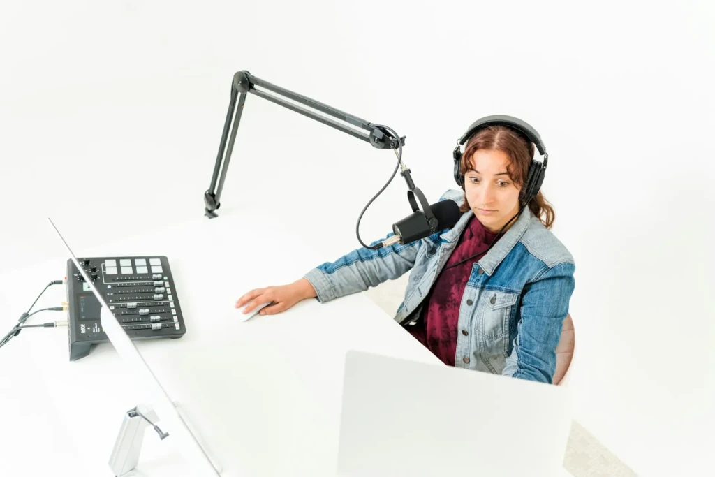 A woman wearing headphones and a denim jacket speaks into a microphone at a desk with audio equipment, conveying focus and professionalism.