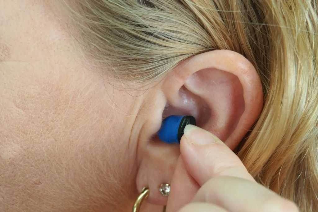 A close-up of a person inserting a blue-and-black custom-molded earplug into their ear canal, with blonde hair pulled back and a gold hoop earring visible, illustrating proper fit and insertion technique for hearing protection or in-ear audio devices.