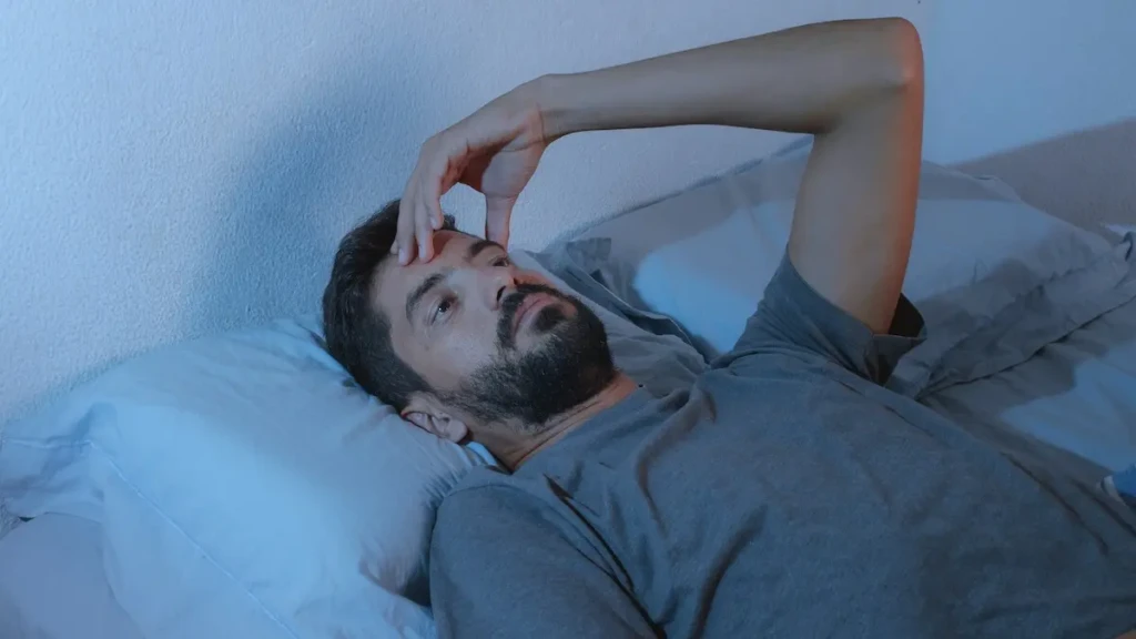 A man with a beard lies awake in bed, looking thoughtful and concerned. The room is dimly lit with a blue hue, conveying a mood of insomnia or worry.