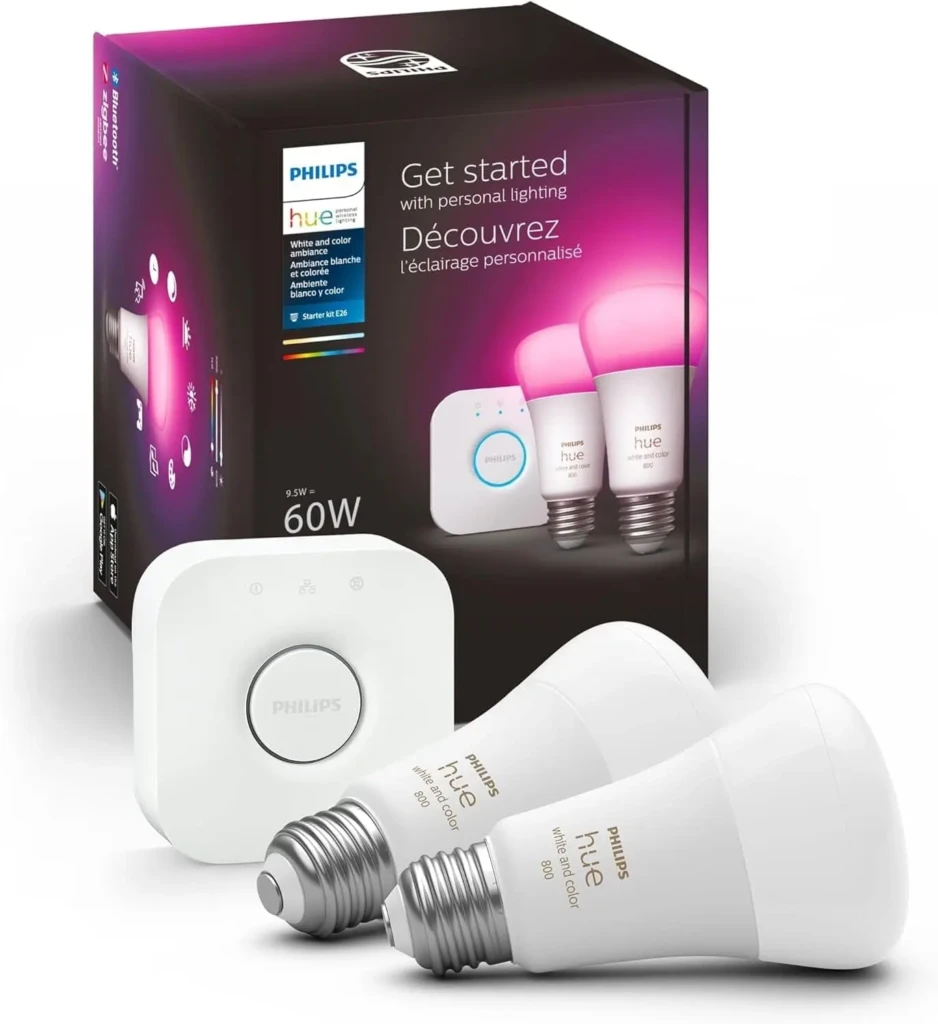 A Philips Hue Starter Kit E26 box with bilingual text (“Get started with personal lighting” / “Découvrez l’éclairage personnalisé”), alongside a white bridge hub and two white “white and color” LED bulbs, illustrating an entry-level smart lighting system for customizable ambiance.
