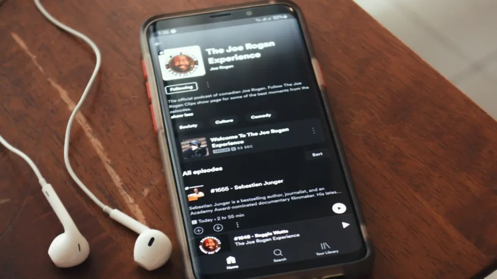 A smartphone on a wooden table displaying the Spotify podcast page for “The Joe Rogan Experience,” with white wired earbuds resting beside it, highlighting podcast discovery and listening on mobile within Spotify’s ecosystem.
