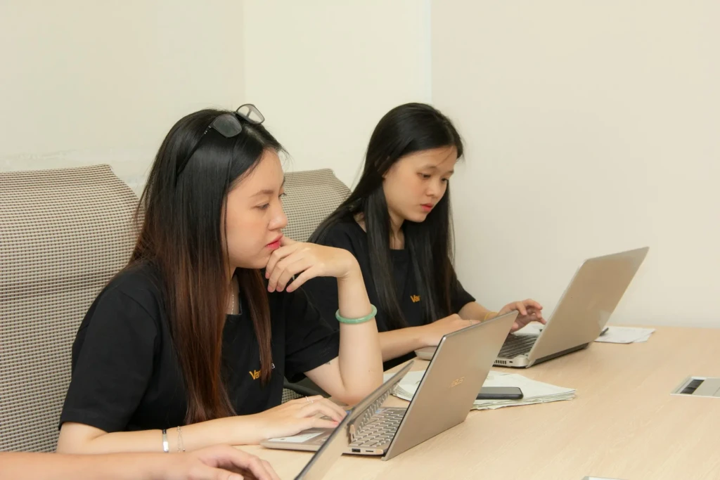 Two women are seated at a wooden table, focused on their ASUS laptops, wearing matching black shirts with a logo; papers and a smartphone rest nearby, depicting collaborative work or professional learning in an office or workshop setting.