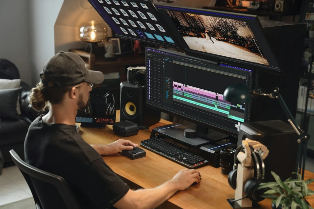 A person from behind, wearing a cap and headphones, editing video on a dual-monitor setup: the main screen shows a timeline in DaVinci Resolve or similar software, while the top screen displays a wedding scene; the desk includes a jog shuttle, camera, speakers, and a lamp, depicting an advanced creator’s editing station with ergonomic and professional tools.