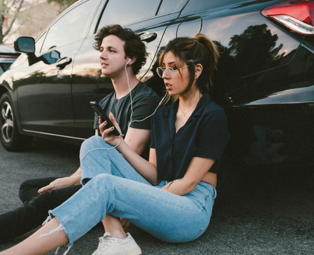 A young man and woman sitting on asphalt beside a black car, sharing white wired earbuds connected to a smartphone the woman holds, evoking casual, shared music listening in everyday life, often associated with streaming service usage.