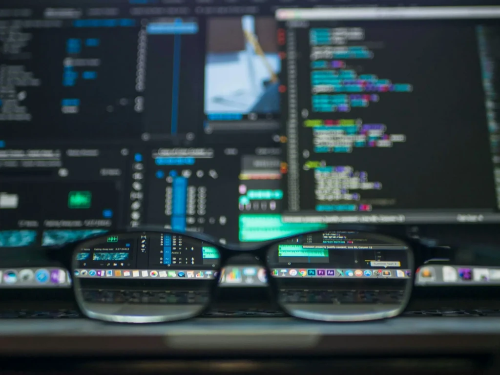 A pair of eyeglasses resting on a desk, with dual monitor screens reflected in the lenses displaying colorful code editors, terminal panels, and UI toolbars, symbolizing a developer’s immersive, multi-tasking workflow in a modern coding environment.