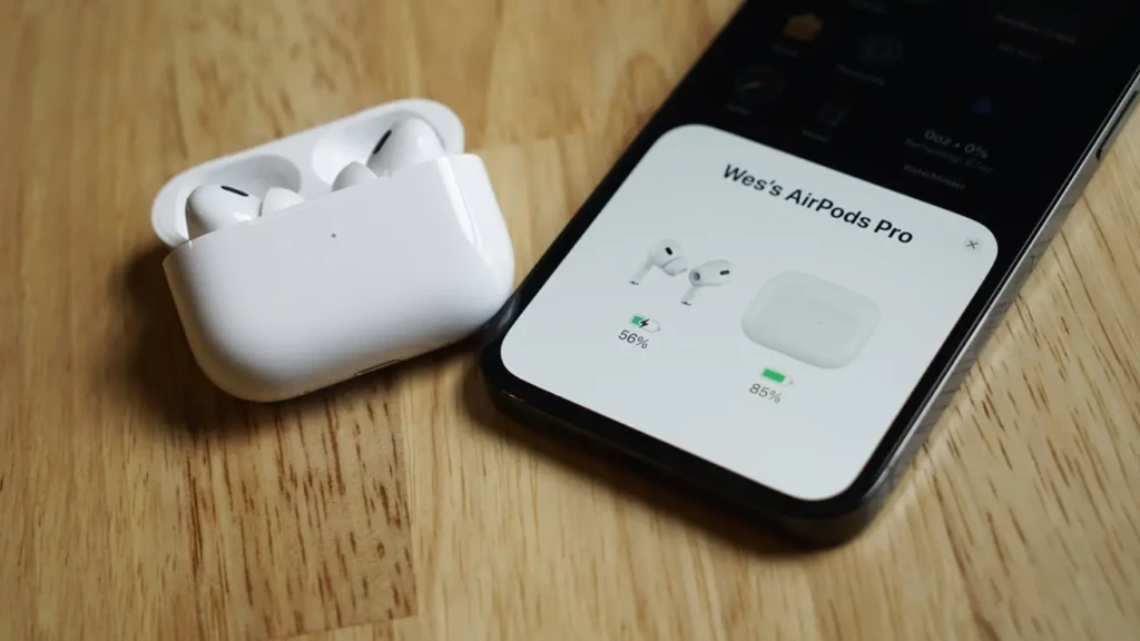 A pair of white AirPods Pro in their open charging case on a wooden table next to a smartphone displaying a battery status screen for the AirPods.