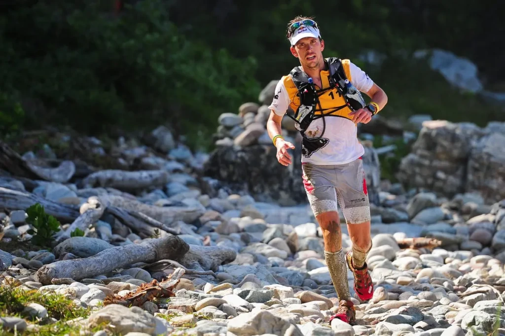 A male trail runner mid-stride on a rocky, uneven terrain, wearing a hydration vest, race bib, and cap, captured in action during an off-road race, exemplifying the rugged, endurance-focused use case often tracked by both NRC and Strava.