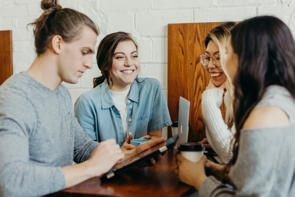 Four people collaborating around a wooden table, smiling and engaged with a tablet, laptop, and coffee cups in a bright, modern café setting, illustrating real-world teamwork and productivity, often associated with digital note-taking and idea-sharing workflows.