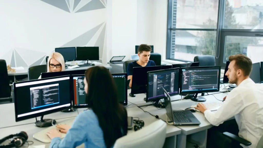 Office scene with four people coding on computers. Two rows of desks, monitors displaying code. Bright, modern space with large windows in the background.
