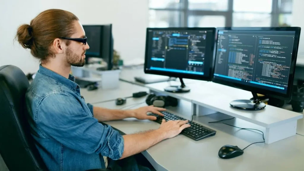 A developer with long hair in a bun and sunglasses sits at a dual-monitor workstation, typing on a keyboard while coding in IDEs displayed on both screens, with headphones and a mouse nearby, illustrating a professional, multi-display software development environment.