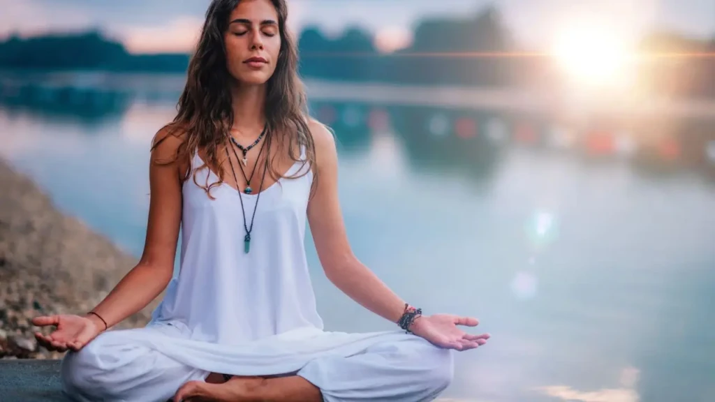 A woman with long hair meditating in lotus position by a calm lakeside at sunset, wearing white clothing and layered necklaces, with eyes closed and palms upturned, conveying mindfulness, tranquility, and connection with nature.
