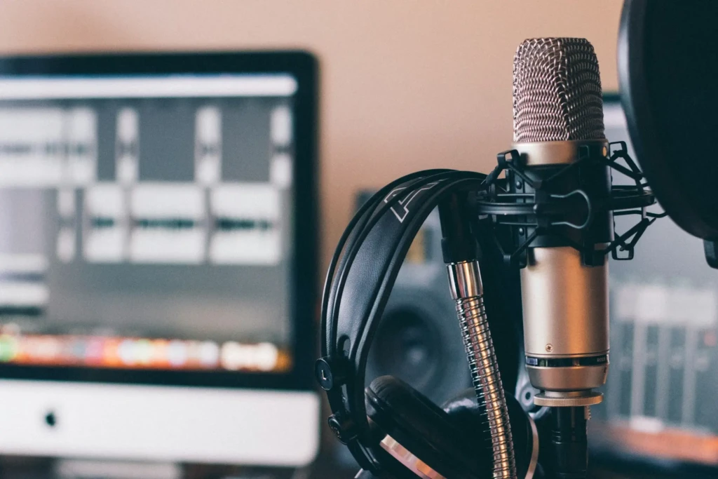 A silver microphone with a pop filter is close to a pair of black headphones, set against a blurred computer screen displaying audio editing software.