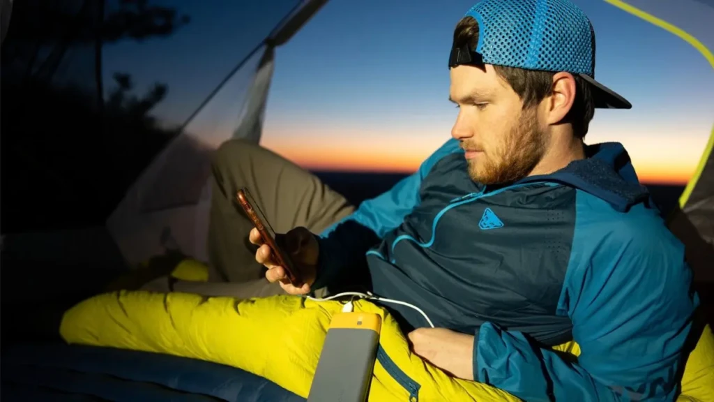 A man inside a tent at dusk using a smartphone powered by a yellow-and-gray portable charger resting on his lap, with a sunset visible through the tent opening, demonstrating real-world mobile charging during travel or off-grid scenarios.