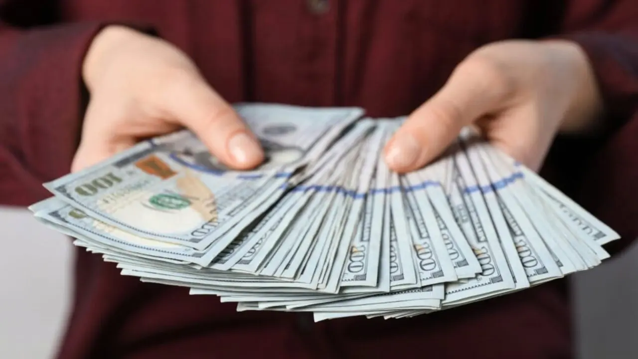 Hands holding a large stack of U.S. hundred-dollar bills, fan-shaped. The person is wearing a dark shirt. The image conveys wealth and abundance.