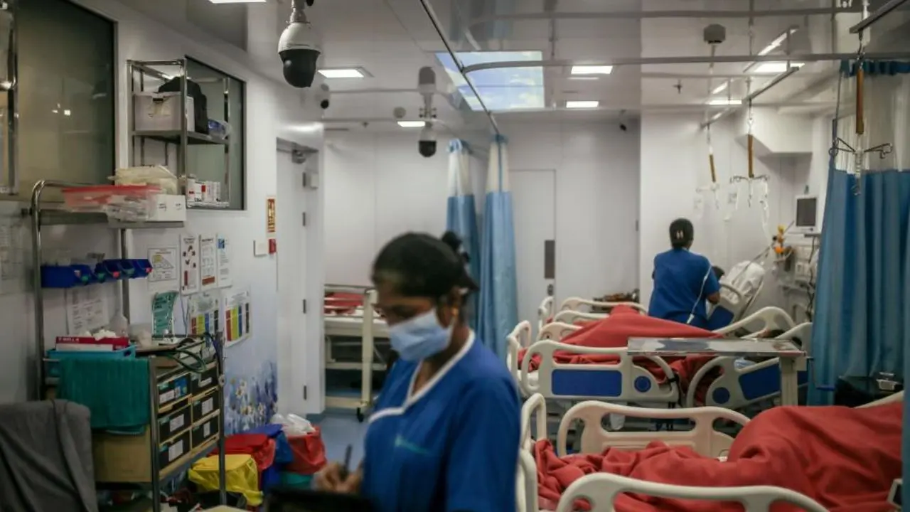 Hospital ward with medical staff tending to patients in bed, covered by red blankets. Monitors and equipment surround the clean, sanitary space.
