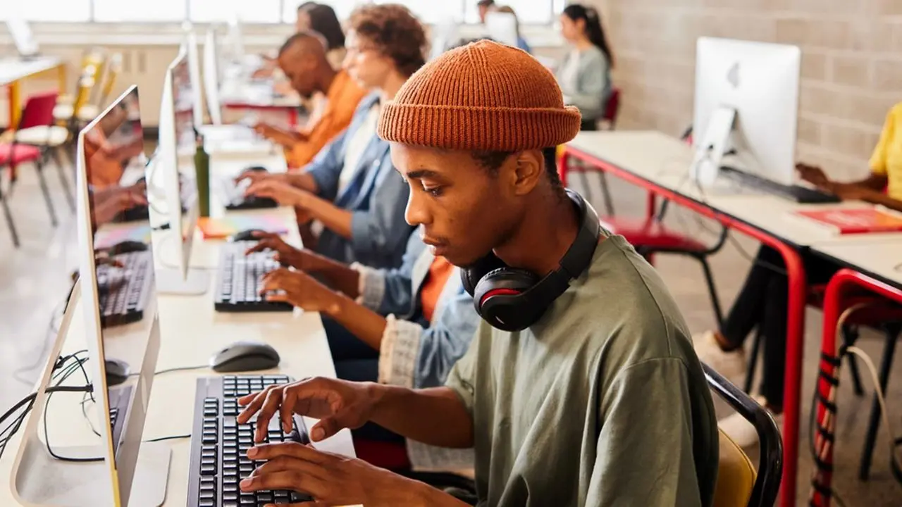 A focused student wearing a beanie and headphones works on a desktop computer in a classroom. Several others are seated at computers in the background.