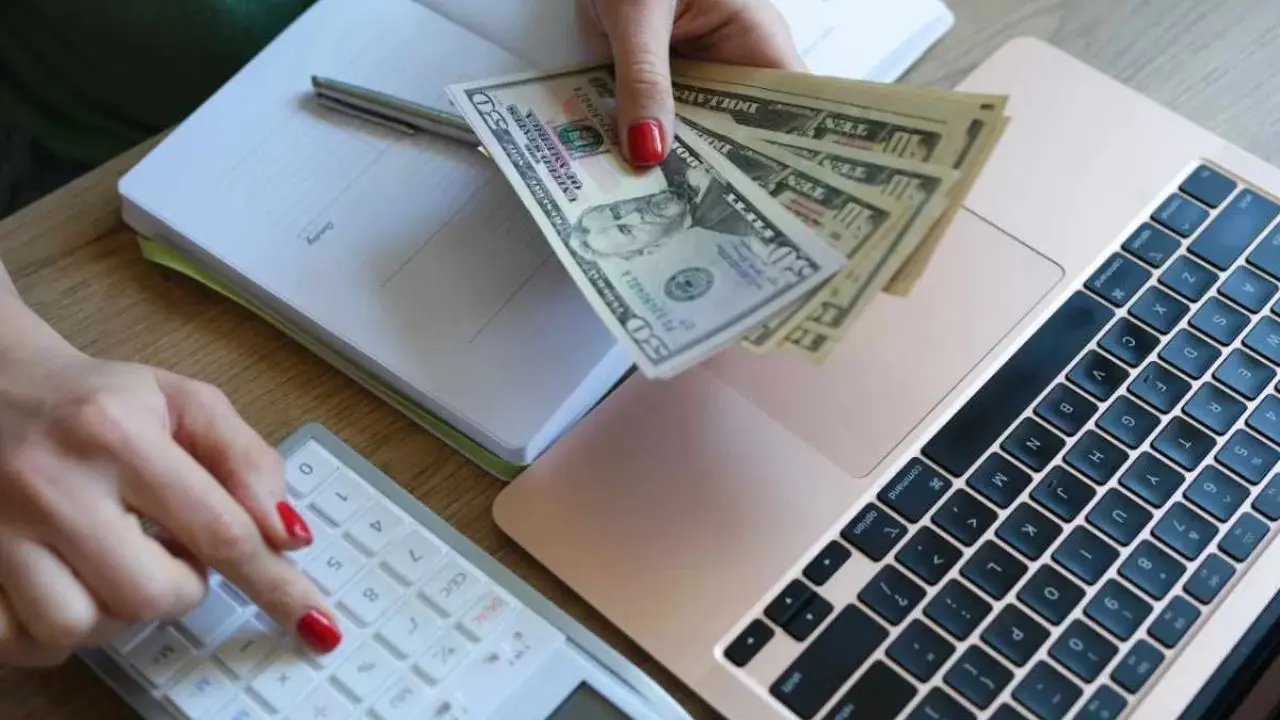 A person with red nails holds dollar bills over a laptop, using a calculator beside an open notebook. The scene conveys financial planning.