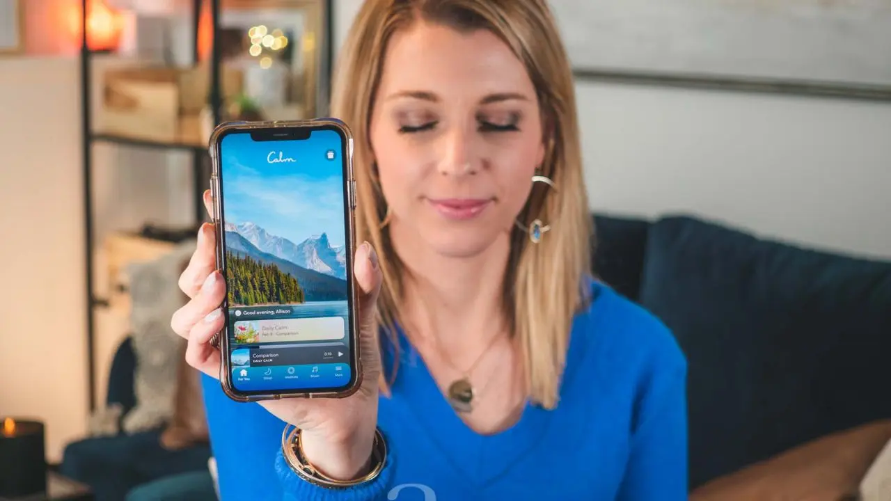 A woman in a blue sweater holds up a smartphone displaying a meditation app. She appears relaxed, with eyes gently closed, in a cozy living room.