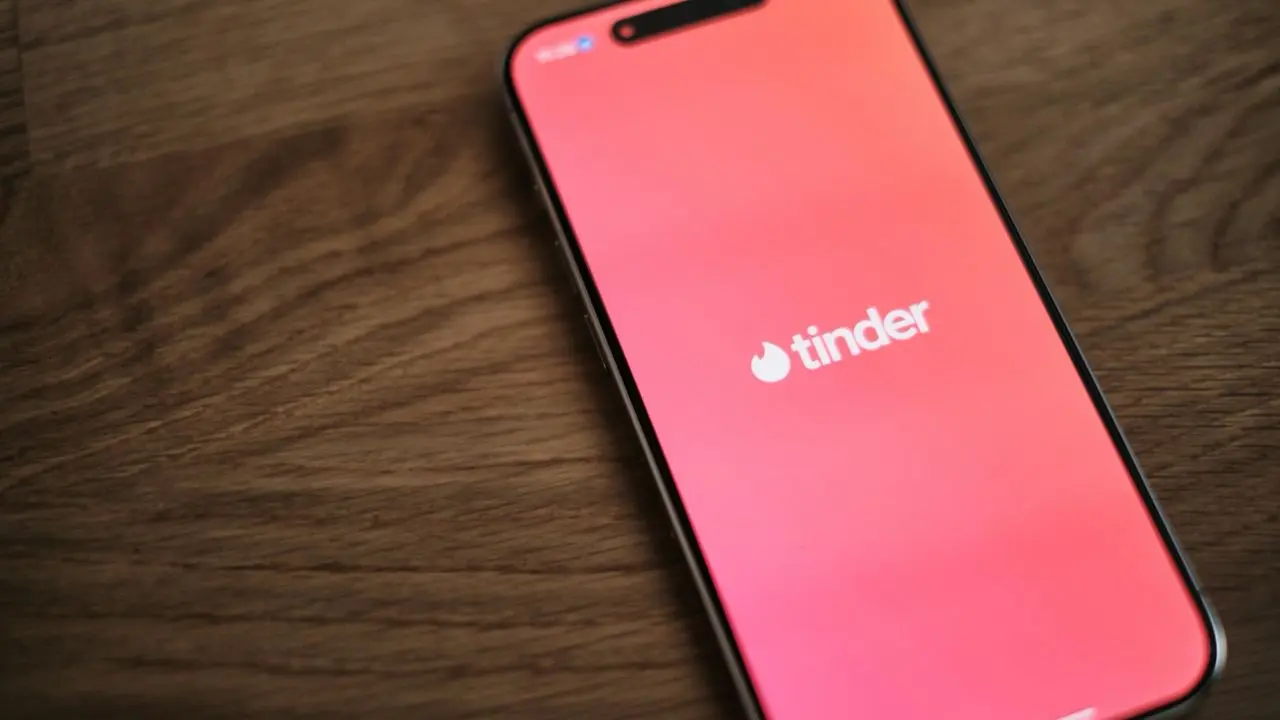 Smartphone displaying the Tinder app on a wooden surface. The screen shows the app's logo on a bright red background, suggesting social connection.