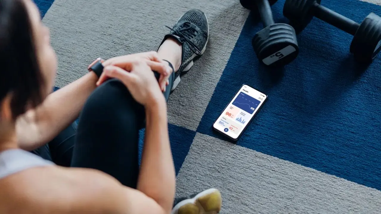 A person sitting on a checkered floor, viewing fitness stats on a smartphone. Nearby are black dumbbells, suggesting a workout setting.