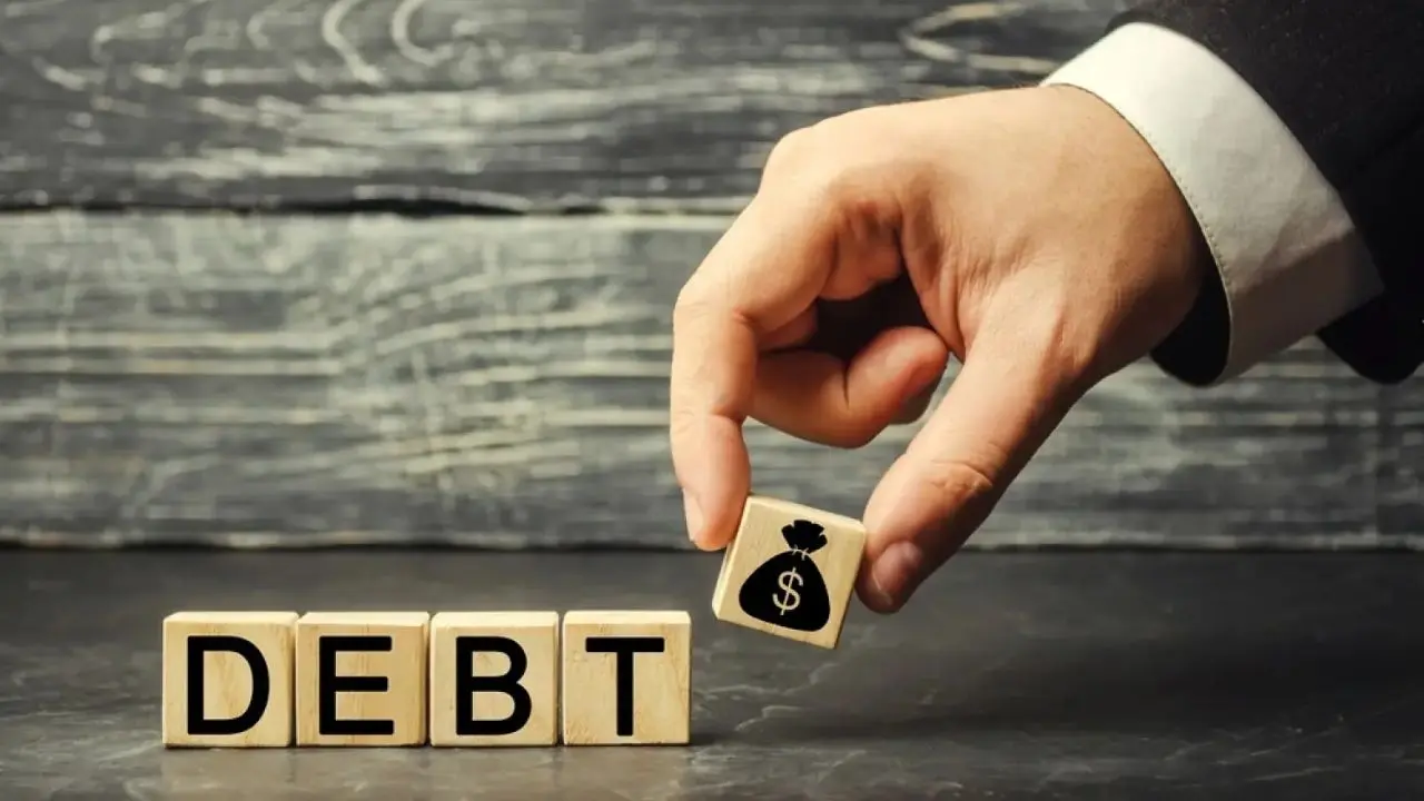 A hand in a suit places a wooden block with a money bag symbol beside blocks spelling "DEBT," suggesting financial burden. Background is textured wood.