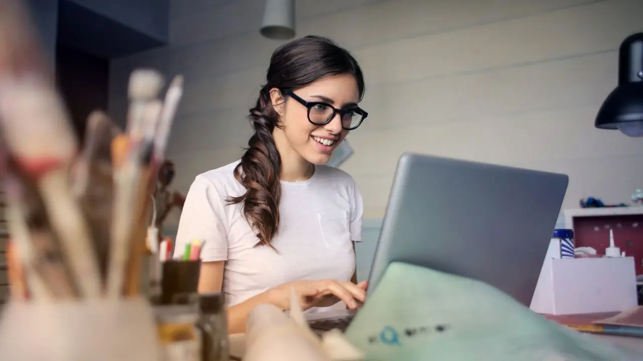 A woman with glasses, smiling, works on a laptop in a creative workspace with art supplies, conveying a focused and joyful atmosphere.