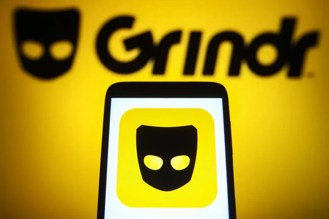 A smartphone displays the Grindr app icon, featuring a stylized black mask on a yellow background. The Grindr logo is prominently visible in the background.