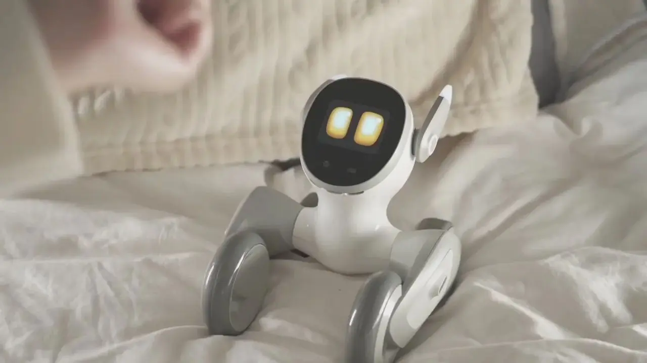 A small, friendly robot sits on a bed, displaying bright eyes on a screen. It has wheels for mobility, conveying a playful and inviting tone.