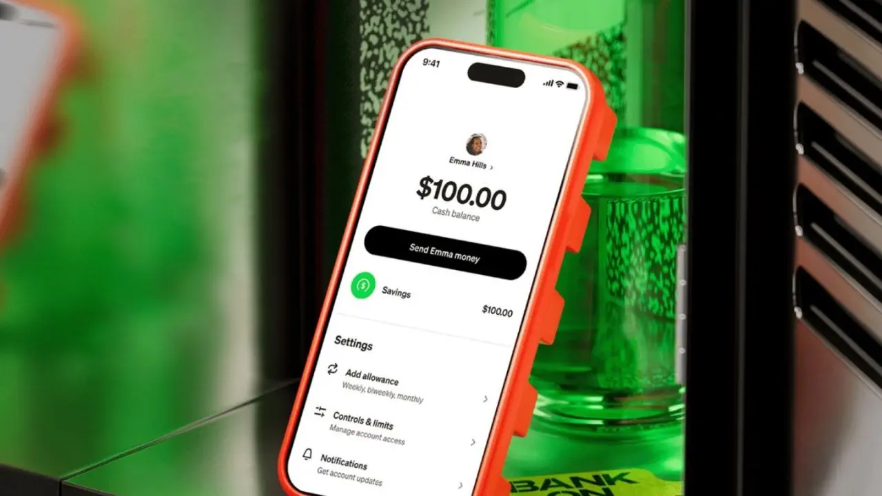 A smartphone with an orange case displays a balance of $100 in a finance app. The phone leans against a green ATM, suggesting mobile banking.