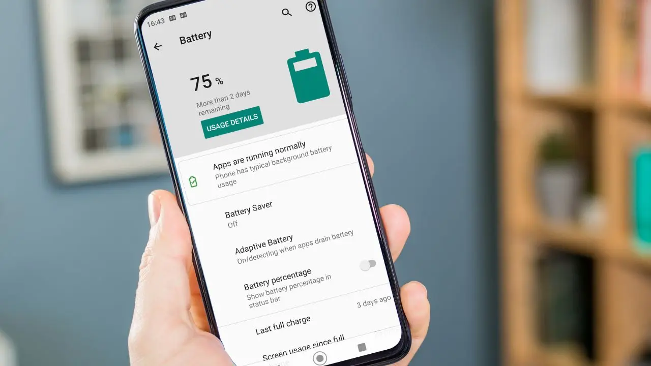 A hand holds a smartphone displaying battery settings, showing 75% charge and over two days remaining. Background is blurred with a softly lit room.