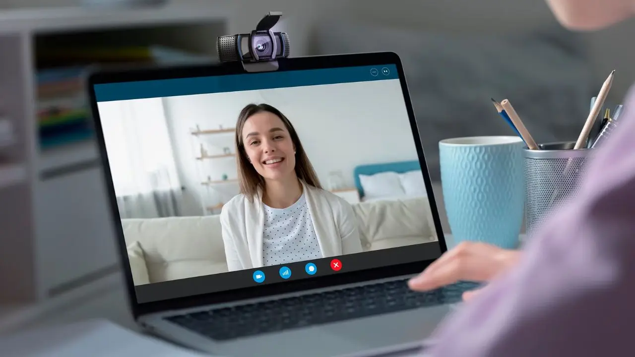 A woman smiling during a video call on a laptop screen. The setting is cozy, with pastel decor. The mood is friendly and relaxed, suggesting a casual conversation.