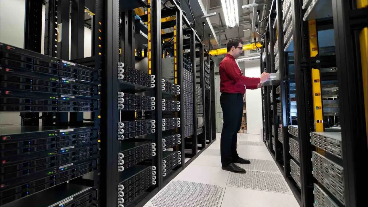 A person in a red shirt works on a laptop among towering, organized server racks in a bright, spacious data center, conveying a sense of focus and technology.